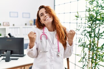 Poster - Young redhead woman wearing doctor uniform and stethoscope at the clinic very happy and excited doing winner gesture with arms raised, smiling and screaming for success. celebration concept.