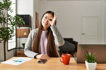 Sticker - Young chinese business worker wearing business style sitting on desk at office smiling happy doing ok sign with hand on eye looking through fingers