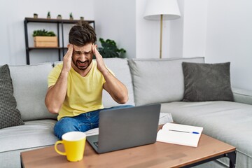 Poster - Young man with beard using laptop at home with hand on head for pain in head because stress. suffering migraine.