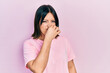 Young hispanic woman wearing casual pink t shirt smelling something stinky and disgusting, intolerable smell, holding breath with fingers on nose. bad smell
