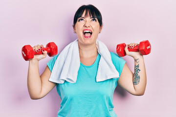 Canvas Print - Young hispanic woman wearing sportswear using dumbbells angry and mad screaming frustrated and furious, shouting with anger looking up.