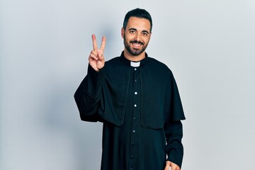 Canvas Print - Handsome hispanic man with beard wearing catholic priest robe smiling looking to the camera showing fingers doing victory sign. number two.