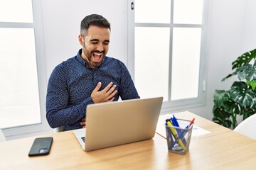 Poster - Young hispanic man with beard working at the office with laptop smiling and laughing hard out loud because funny crazy joke with hands on body.