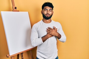 Sticker - Arab man with beard standing by painter easel stand smiling with hands on chest, eyes closed with grateful gesture on face. health concept.