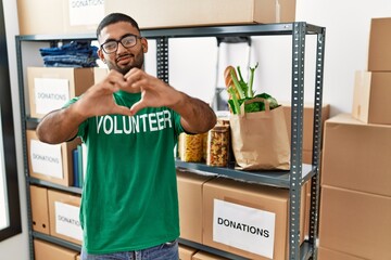 Poster - Young indian man volunteer holding donations box smiling in love doing heart symbol shape with hands. romantic concept.