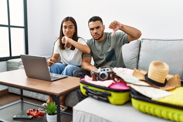 Canvas Print - Young interracial couple packing summer clothes in suitcase looking at laptop with angry face, negative sign showing dislike with thumbs down, rejection concept