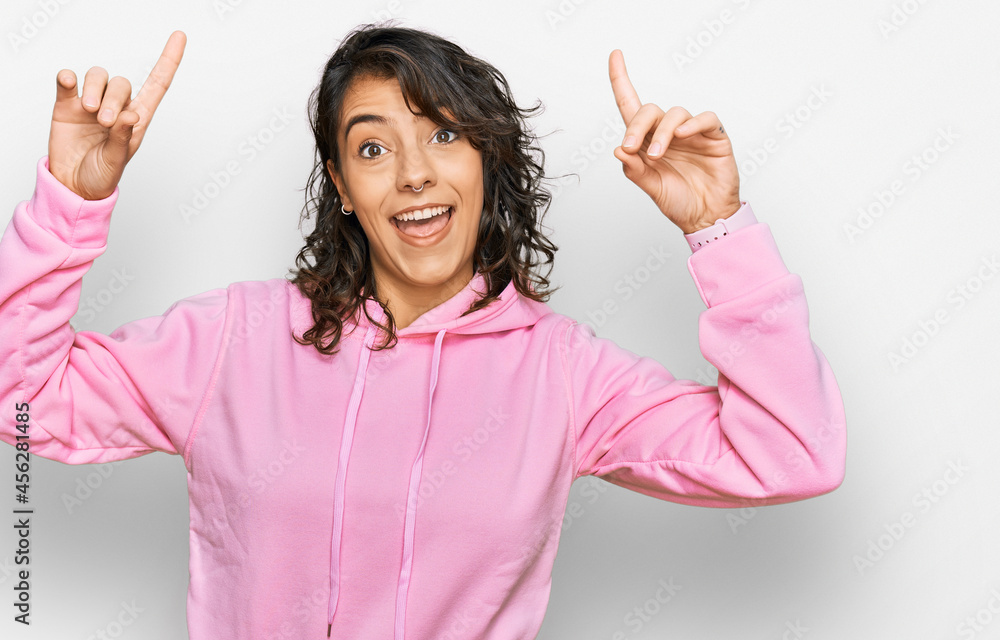 Young hispanic woman wearing casual sweatshirt smiling amazed and surprised and pointing up with fingers and raised arms.