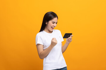 Excited Beautiful young asian women play mobile game and standing on isolated yellow background. Playing game on smartphone winning victory moment. Very enjoy and fun relax time