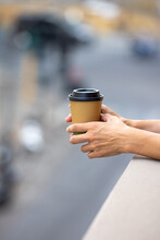 Hands Holding A Single Use Paper Coffee Cup. Young Woman In A Balcony Drinking Coffee From Disposable Cup With A Blurred City View.  