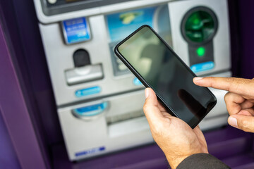 Young businessman standing front of atm machine using smartphone withdraw the money without atm credit card. He ut the pin code into smartphone and take the money.