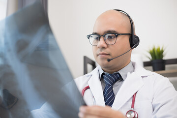 Doctor talks to patient via online video conferencing with computer wearing headset. Explain about taking medications to treat illnesses holding x-ray film.