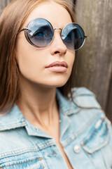 Wall Mural - Fashion portrait of beautiful woman with caucasian face in fashionable round sunglasses with vintage jeans jacket outdoors