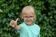 A small blonde girl with glasses gives a thumbs up gesture in a mint T-shirt on a green background, a happy child smiles