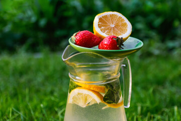 Defocus saucer with lemon and two strawberry standing on glass jug of lemonade with slice lemon and leaves of mint on nature green background. Pitcher of summer cocktail. Out of focus