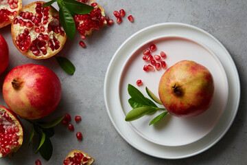 Wall Mural - Ripe pomegranate fruit and half, seeds pomegranate with leaves on grey concrete background. Fresh pomegranate on white plate. Vegan, healthy diet concept. Top view. 