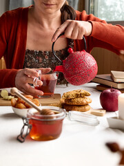 Woman pours a hot tea from red teapot in a glass mug on the table. Honey, biscuits and books near the cup of tea on the linen tablecloth. Aromatic mood. Cozy fall vibes. Tea drinking.Selective focus.