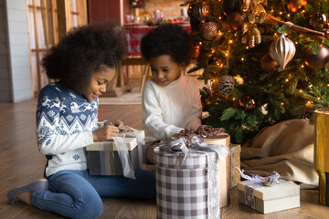 Wall Mural - Happy small teen African American children sit on floor near fir tree open gifts presents on Christmas morning. Smiling little biracial kids enjoy winter holidays unpack boxes for New Year.