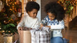 Happy small African American kids feel excited open unbox parcels with gifts presents for New Year. Smiling little teen children unpack box celebrate Christmas together at home. Celebration concept.