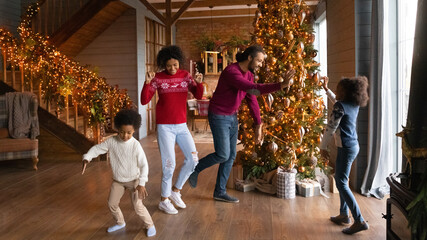 Wall Mural - Happy young African American family with kids dance near fir tree in cozy home relax on New Year winter holidays together. Smiling biracial parents with children rest on Christmas play and jump.