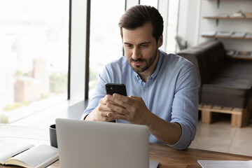 Wall Mural - serious young caucasian male employee sit at desk in office work on computer browse internet on cell