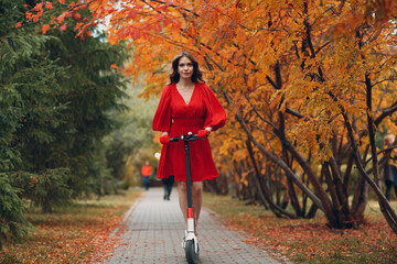 Wall Mural - Young woman with electric scooter in red dress at the autumn city park.