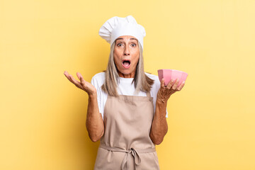 middle age chef woman feeling extremely shocked and surprised, anxious and panicking, with a stressed and horrified look and holding an empty pot