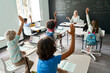 © insta_photos - Happy Caucasian female teacher having maths lesson with elementary diverse schoolchildren sitting at desk using laptops. Full class of kids raising hands knowing answer to task written on chalkboard.