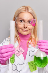 Experienced woman professor works on new scientific research compares liquid in flask and tube wears transparent eyeglasses rubber gloves and white coat poses in laboratory against grey background