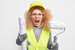 Emotional curly female painter holds rolled paper blueprint and roller works on new architectural project renovates apartment wears hardhat uniform safety glasses isolated over white background