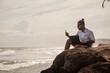 Cyfrowy nomada, człowiek pracujący z laptopem i smartfonem zdalnie na tle oceanu i skał siedzący na wybrzeżu.