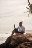 Fototapeta  - Cyfrowy nomada, człowiek pracujący z laptopem i smartfonem zdalnie na tle oceanu i skał siedzący na wybrzeżu.