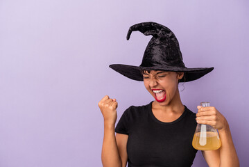 Young mixed race woman disguised as a witch holding potion isolated on purple background  raising fist after a victory, winner concept.