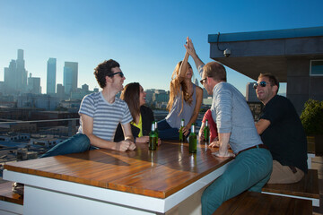 Six adult friends drinking beer at table of rooftop bar with Los Angeles skyline, USA