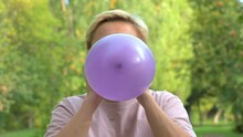 A Blonde Girl With Short Hair Inflates A Balloon Against The Background Of Green Nature Close-up