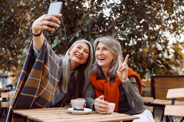 Wall Mural - Joyful senior women friends take selfie with modern smartphone sitting at small table in street cafe on nice autumn day