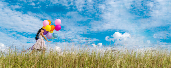 Wall Mural - Banner Cheerful cute girl holding balloons running green meadow white cloud and blue sky with happiness. Panorama Hands holding vibrant air balloons happy times summer sunlight outdoor copy space