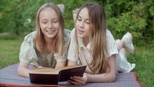 Happy Young Girls Lying And Reading Book In Park. Cheerful Teenage Friends Lying Together On Green Grass, Laughing And Talking About Book At Summertime