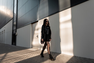 Fashion girl model with sunglasses in trendy black clothes with leather jacket and handbag in sexy tights with shoes walks on the street near a modern building at sun light. Urban female style