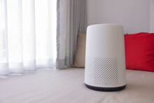 White Air Purifier Sits In The Living Room With White Curtains And Red-brown Pillows In The Background. Protect PM 2.5 Dust And Air Pollution Concept. Air Cleaner Removing Fine Dust In House