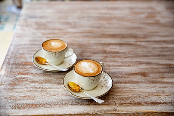 Two stylish white cups of cappuccino with latte art on saucer and golden spoons on light wooden table.