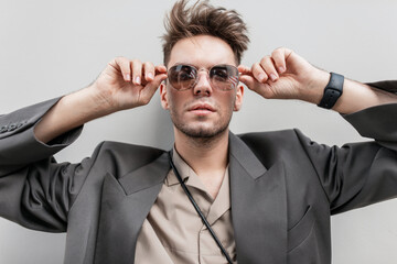 Wall Mural - fashion portrait of a handsome man model with stubble in fashionable casual outfit with a blazer and shirt looks through sunglasses near a gray wall