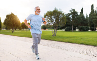 Happy senior man in headphones listening music while jogging outside in city park