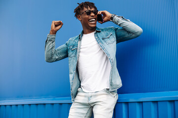 successful american black guy calls on the phone, shows a sign of success and joy to victory, on a blue background