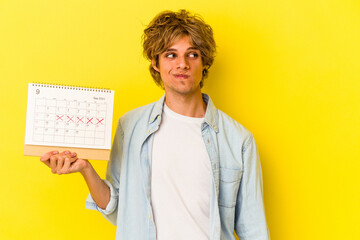 Wall Mural - Young caucasian man with makeup holding calendar isolated on yellow background  confused, feels doubtful and unsure.