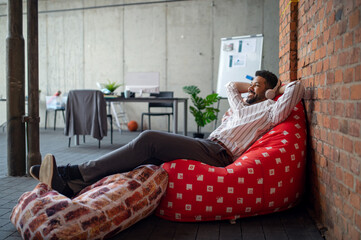 Wall Mural - Cheerful young businessman with headphones lying on bean bag in office, taking a break and relaxing.