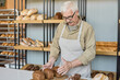 © pressmaster - Serious mature baker in glasses and apron standing at counter and adjusting loaves of brown bread on wooden board