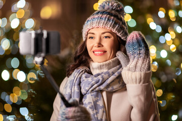 Poster - holidays, communication and people concept - portrait of beautiful happy smiling young woman with smartphone and selfie stick taking picture over christmas lights in winter city