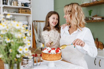 Happy smiling chef cook baker mom woman in white shirt work with child baby girl helper decorate pie with cream at kitchen table home Cooking food concept Mommy little kid daughter prepare fruit cake