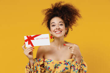 Wall Mural - Young happy satisfied smiling woman 20s with culry hair wear casual clothes hold point finger on gift certificate coupon voucher card for store isolated on plain yellow background studio portrait.