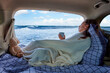 A young woman sitting in a car with the truck open, covered in a cozy blanket while contemplating the ocean view.
